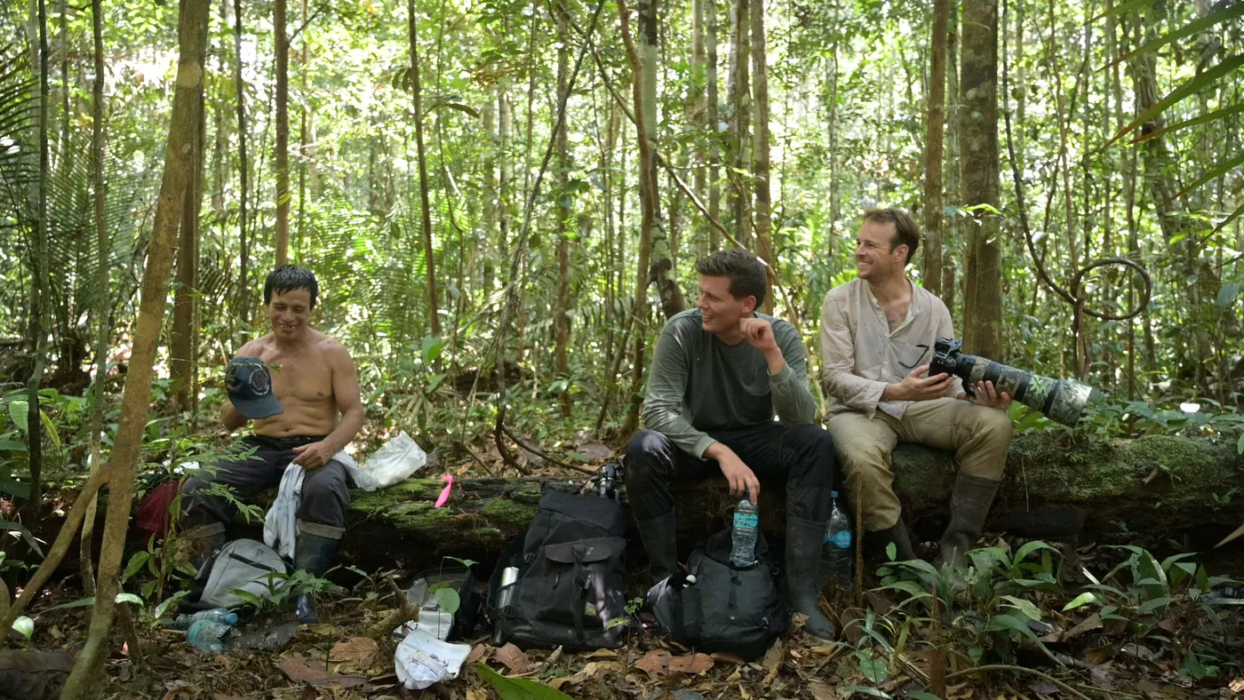 Trois hommes font une pause assis sur un tronc, au milieu de la forêt, avec leur matériel photo.